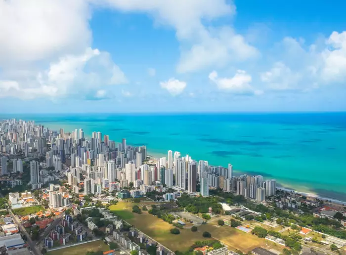 Vista aérea da praia de Boa Viagem em Recife, com prédios e o mar se encontrando em um dia ensolarado.