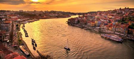 Explore o encanto do pôr do sol dourado de Porto, refletindo no Douro com barcos navegando e a cidade histórica ao fundo.
