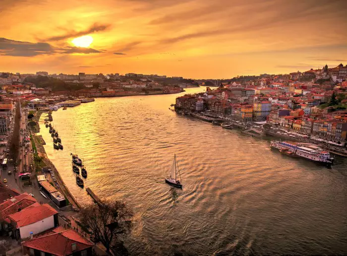 Explore o encanto do pôr do sol dourado de Porto, refletindo no Douro com barcos navegando e a cidade histórica ao fundo.