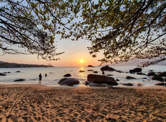 Lindo pôr do sol de Ilhabela, visto da areia da praia para o mar, com pedras sendo escaladas por turistas.