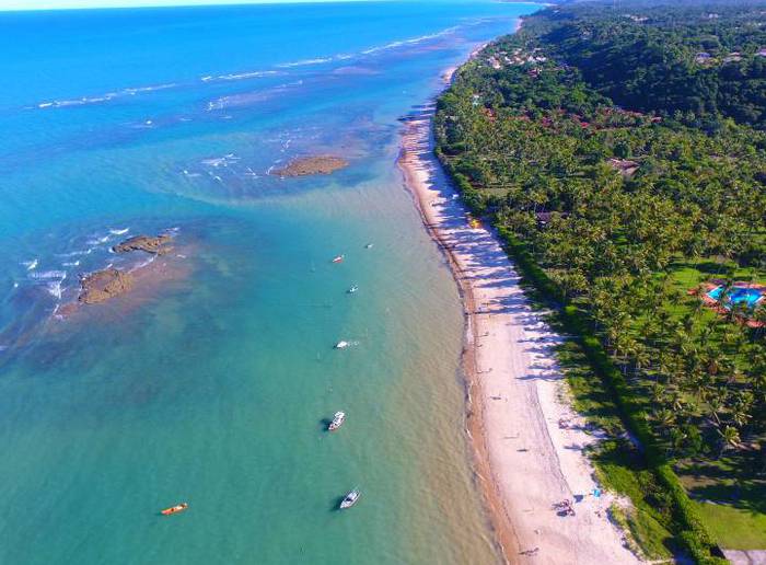 Vista aérea da orla de Arraial d'Ajuda, Bahia. Mar azul e cristalino a esquerda, faixa de areia e vegetação local a direita.