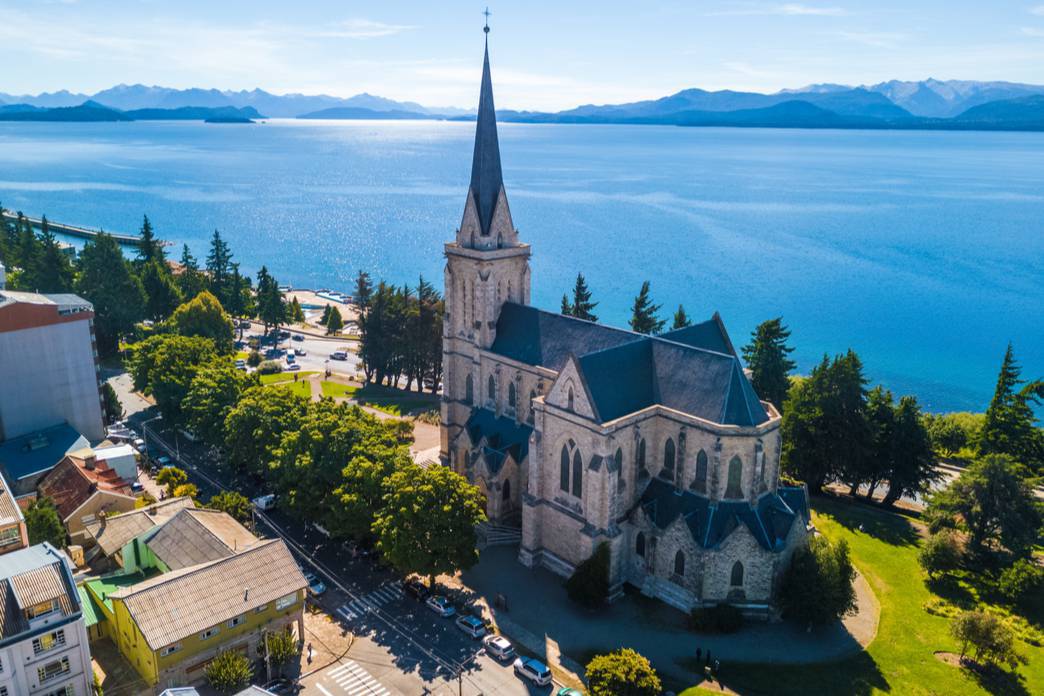 A Igreja de San Carlos de Bariloche se destaca, com sua arquitetura rústica e robusta, harmoniosamente enquadrada pelo céu azul e as montanhas ao fundo.