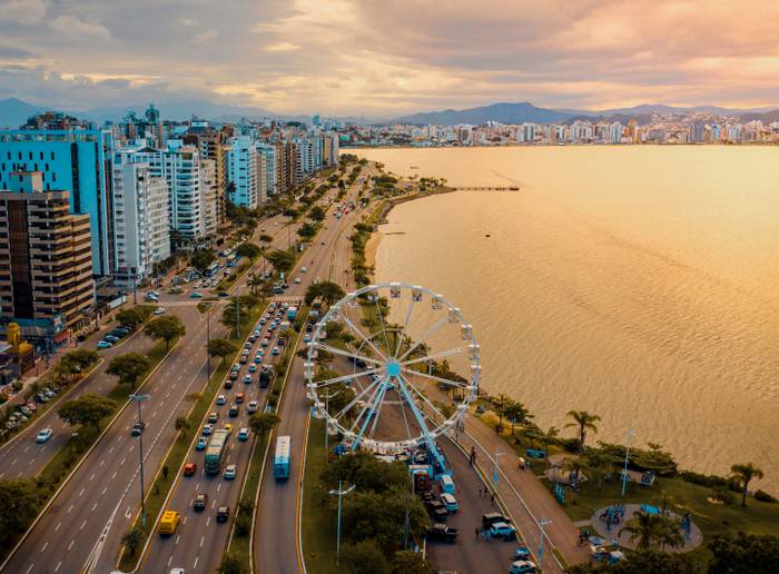 Vista aérea da cidade de Florianópolis à beira-mar, pôr do sol e roda gigante ao fundo.