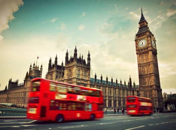 Torre do relógio Big Ben ao fundo da foto, construção na mesma região da Abadia de Westminster. Ônibus vermelho de dois andares passando pela rua, um cartão postal famoso de Londres.