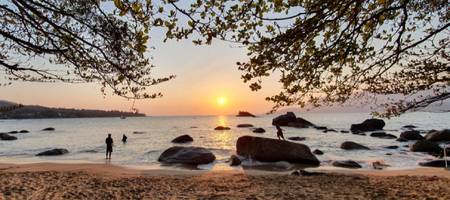 Lindo pôr do sol de Ilhabela, visto da areia da praia para o mar, com pedras sendo escaladas por turistas.
