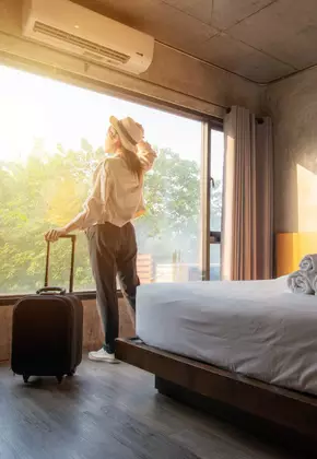 Mulher de chapéu e mala pronta na grande janela de um quarto de hotel com cama arrumada